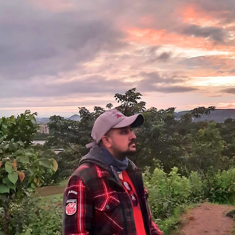 A man in a cap and plaid jacket stands on a dirt path surrounded by greenery, looking ahead as the sun sets behind clouds, casting an orange and pink glow in the sky.